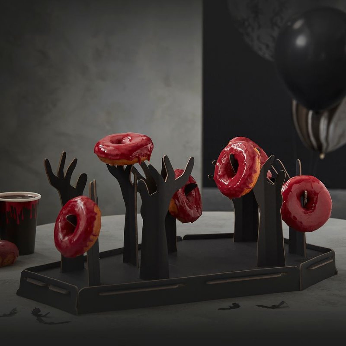 Sort kisteformet donutholder med otte opstrakte zombiehænder, der hver holder en donut med rød glasur, foran en grå væg med sorte balloner