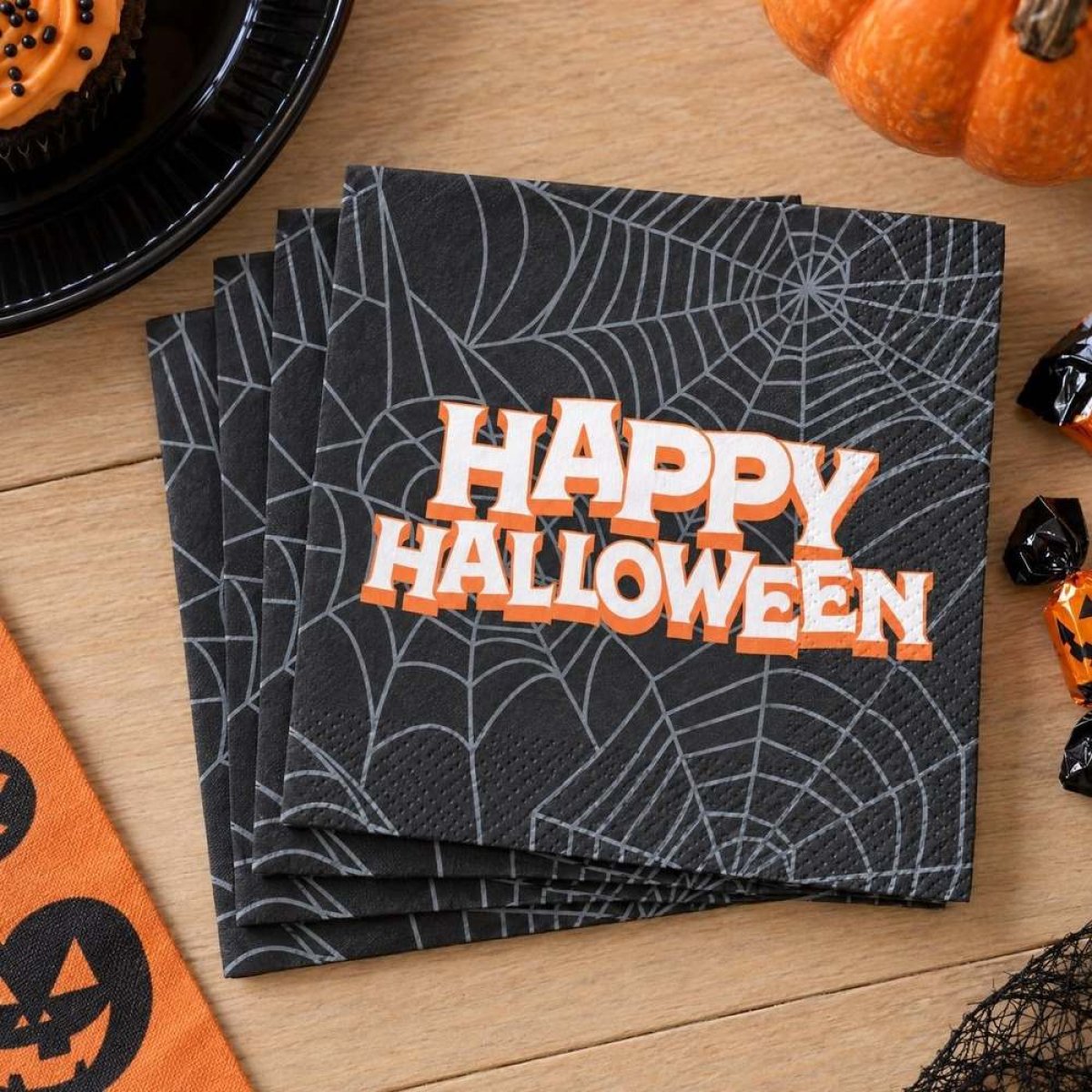 Sorte servietter med teksten Happy Halloween i orange og hvide kanter, lagt i en bunke på et bord med græskar